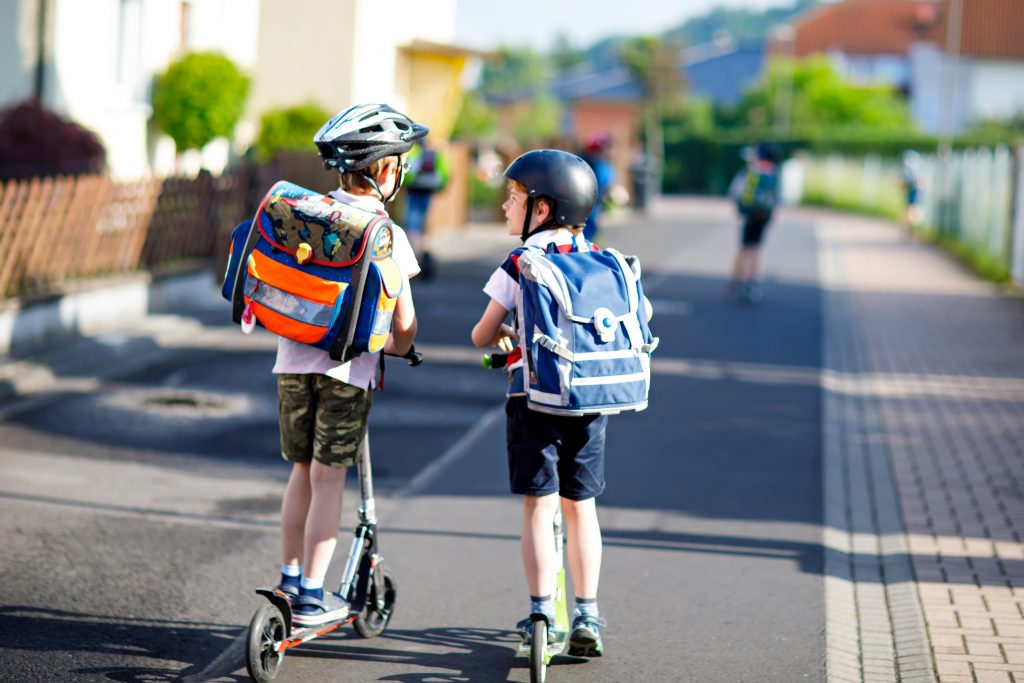 Zwei Schulkind Jungen in Schutzhelm fahren mit Roller in der Stadt mit Rucksack an einem sonnigen Tag.