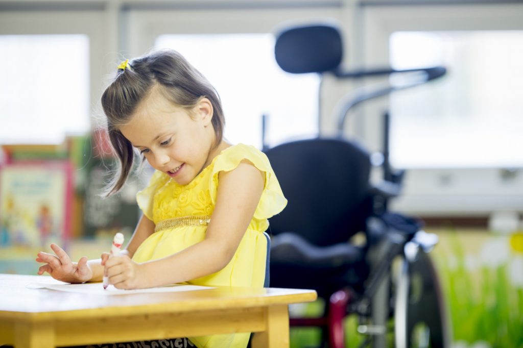 Ein fröhliches kleines Mädchen mit zerebraler Lähmung sitzt an einem Schreibtisch in einer Grundschule und malt ein Bild mit einem Marker aus.